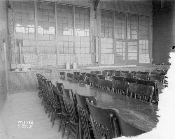 Interior view of rows of tables and chairs in the dining room. Along the back wall are utensils, tableware and urns on top of cabinets, and above are windows.