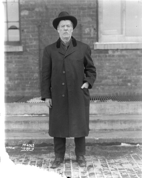 Full-length portrait of a man dressed in business coat standing outdoors. A brick building is behind him.