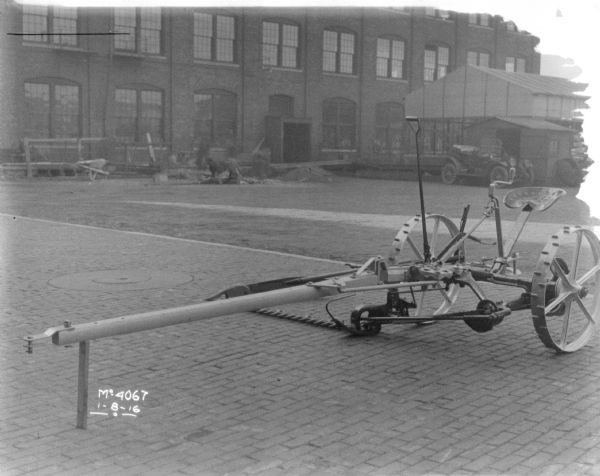 A mower is set-up on the cobblestones outdoors in the factory yard. Outbuildings, an automobile and a brick factory building are in the background.