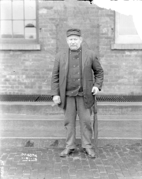 Full-length portrait of a man dressed in a business coat standing outdoors. A brick building is behind him.