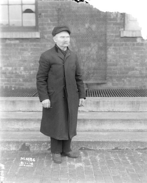 Full-length portrait of a man dressed in warm clothes standing outdoors. A brick building is behind him.
