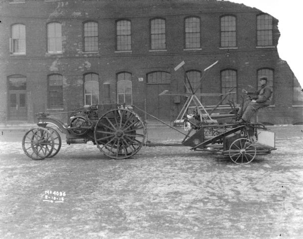 A man is driving a tractor drawn Binder in the factory yard. A brick factory building is in the background.