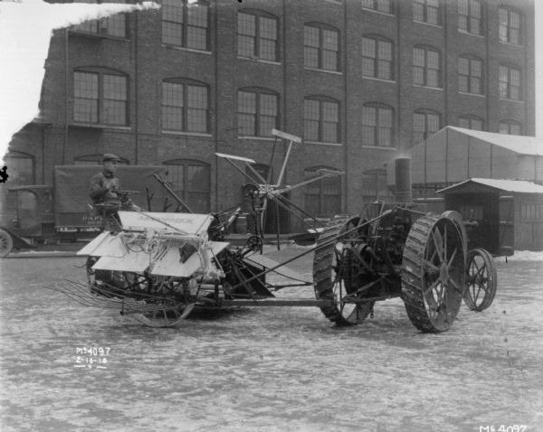 A man is driving a tractor drawn Binder in the factory yard. A truck and outbuildings and a brick factory building are in the background.