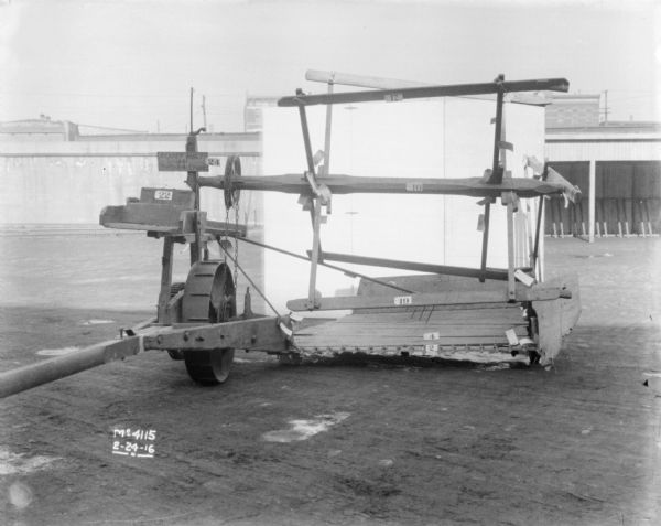 A Reaper is set-up in the factory yard in front of a small backdrop. The Reaper parts are numbered, and a sign on it reads: "Reaper Built 1851 Cut 24 Crops." In the background is an open-sided shed near a fence, and beyond are buildings.