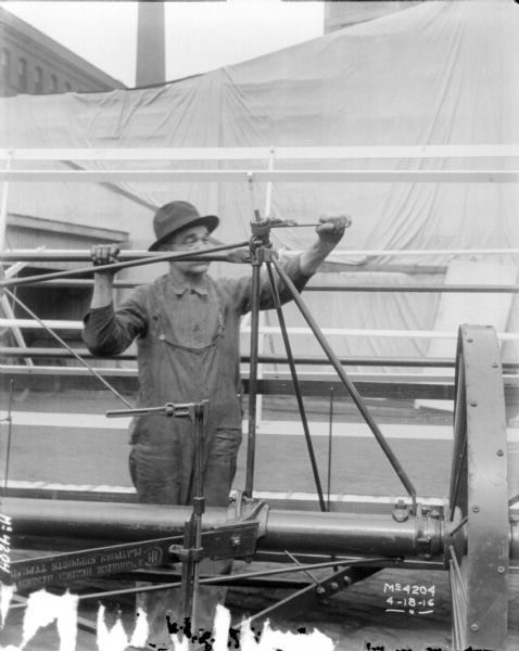 A man is making adjustments on the spring of a Binder outdoors at McCormick Works. In the background is a fabric backdrop, and in the background above is a smokestack and brick building.