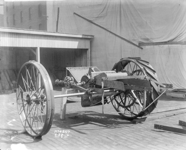 Harvester Thresher parts, including wheels, set-up in factory yard. In the background is bicycle inside an open-sided shed, a backdrop, and brick buildings.