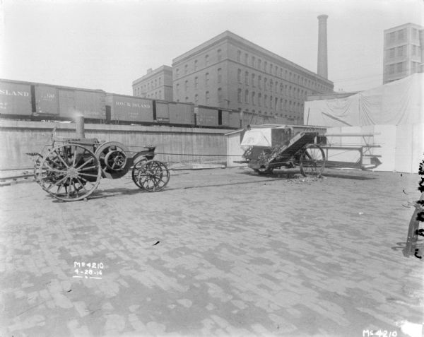 Tractor Powered Harvester Thresher in factory yard. In the background are railroad cars on an elevated platform. A backdrop is set-up on the far right. Brick buildings and a smokestack are in the far background.