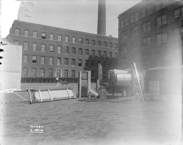 Harvester Thresher packaged for shipping in factory yard. Parts are stacked along the base of a brick building in the background.