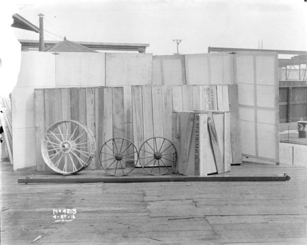 Harvester Thresher parts, including wheels, in factory yard. Some of the parts are packaged in crates for shipping. There is a backdrop behind the parts.