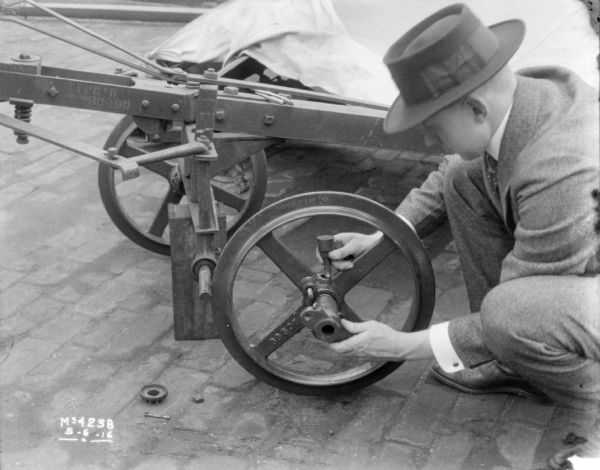Close-up of a man wearing a suit and hat mounting a wheel onto a machine in the factory yard.