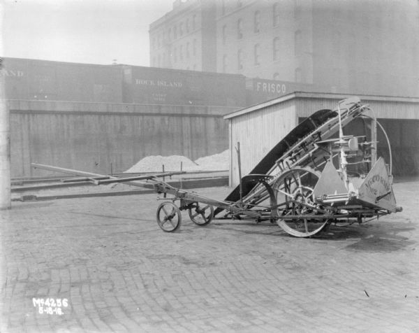 Corn Binder outdoors in the factory yard. In the background is an open sided shed, and railroad cars on an elevated platform. In the far background is a tall, brick building.