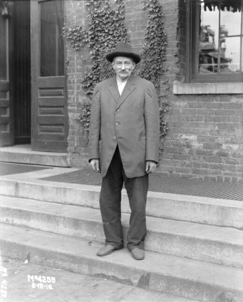 Elderly man, in business suit, smiling and posing on stairs near an entrance to a brick factory building.