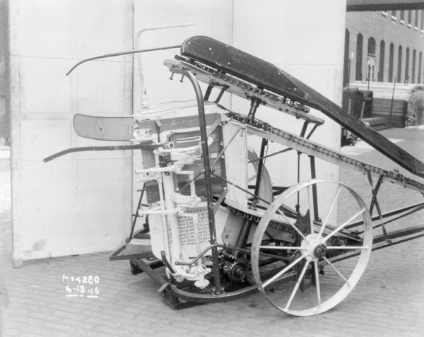 Corn Binder in front of backdrop outdoors in factory yard. A man is working near stacks of material along the side of a brick factory building on the right.