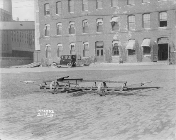 Sweep Rake outdoors in factory yard. In the background an automobile parked near a factory building. An elevated railroad platform is on the left running between the buildings. A skybridge is over the platform.