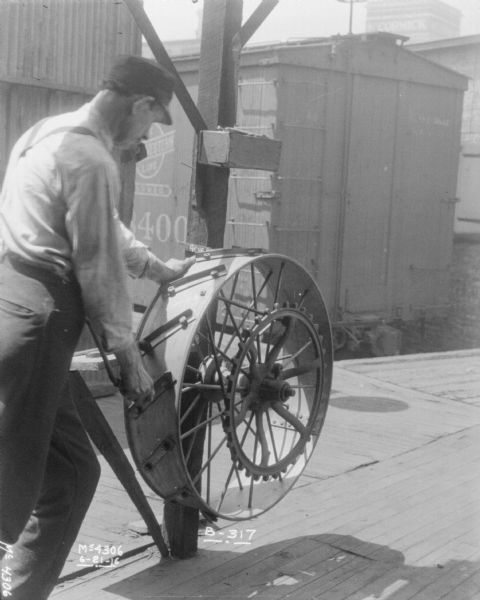 A man is standing outdoors under an open-sided building assembling a wheel on a wooden column. A railroad car and a building are in the background. In the distance is a tall building with a sign near the roof that reads: "McCormick."