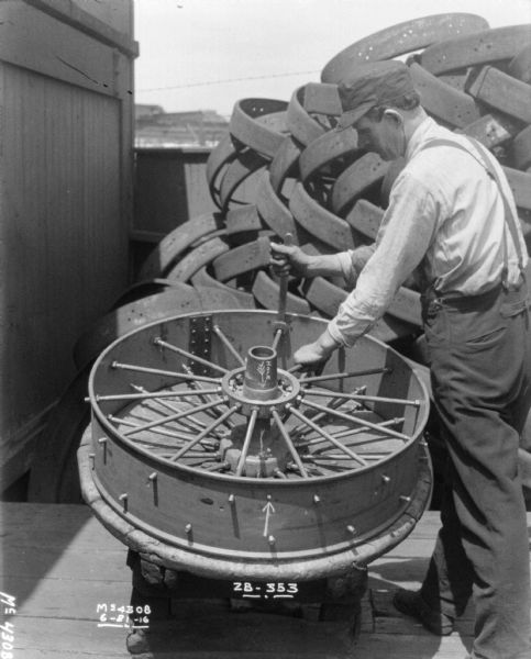 A man is standing outdoors assembling a wheel resting on a round platform. Written under the wheel is "ZB-353." Near the center of the wheel is an arrow, and written alongside is: "Hole." Other parts are stacked on the right, and on the left is a building.