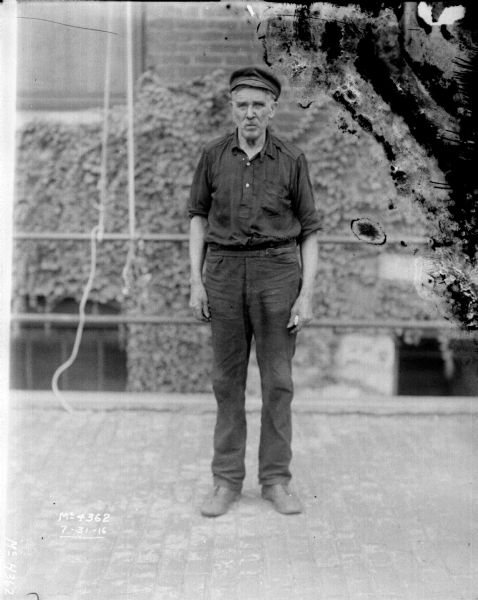Full-length portrait of a man standing outdoors in the factory yard wearing work clothes and a hat. A brick building is in the background.