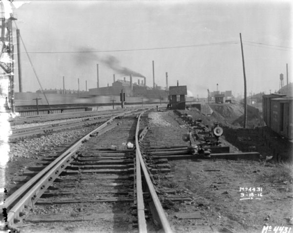 View down railroad tracks in a railroad yard. Factory buildings are in the distance. A man is sitting near a small building near the tracks.