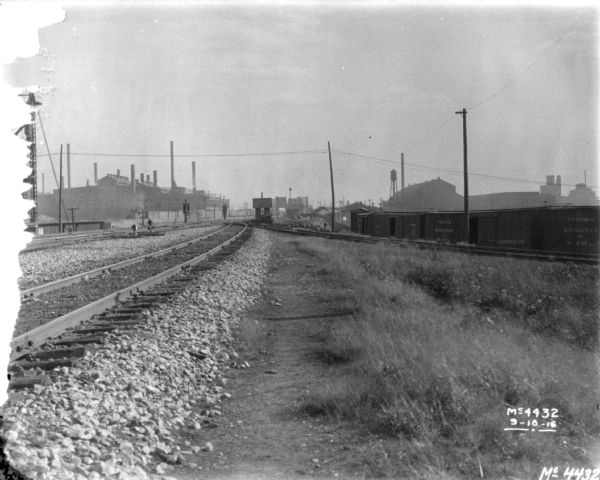 View down railroad tracks in a railroad yard. On the right railroad cars are on another set of tracks. Factory buildings are in the distance.