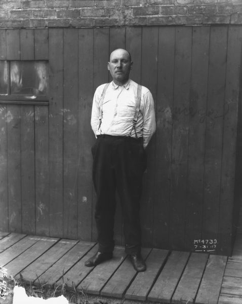 Full-length portrait of a man wearing work clothes standing on a wooden sidewalk in front of a wood wall, with a brick wall above.