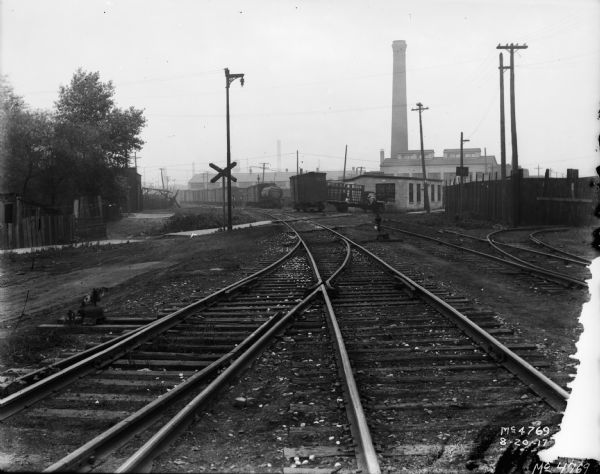 View down sets of railroad tracks towards a railroad crossing and factory buildings. There are smokestacks and railroad cars in the distance.