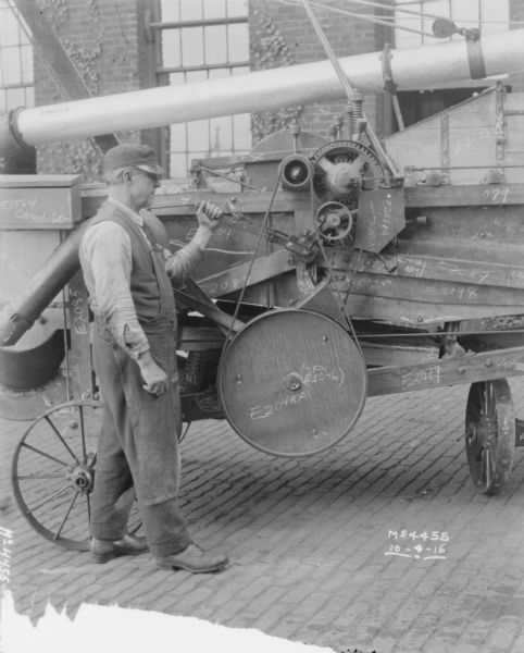 A man is standing and holding onto a part of a Thresher outdoors in the factory yard. Part numbers are written in chalk on the machine. A brick building is in the background.