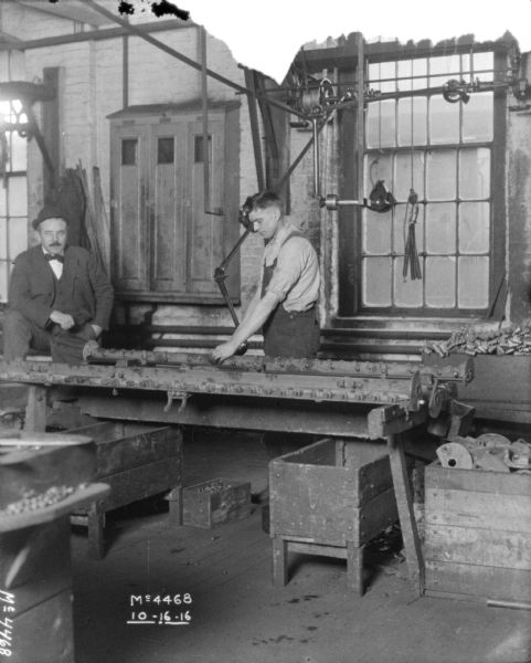 A man is riveting pieces indoors at McCormick Works. Another man wearing a hat and suit is standing on the left.