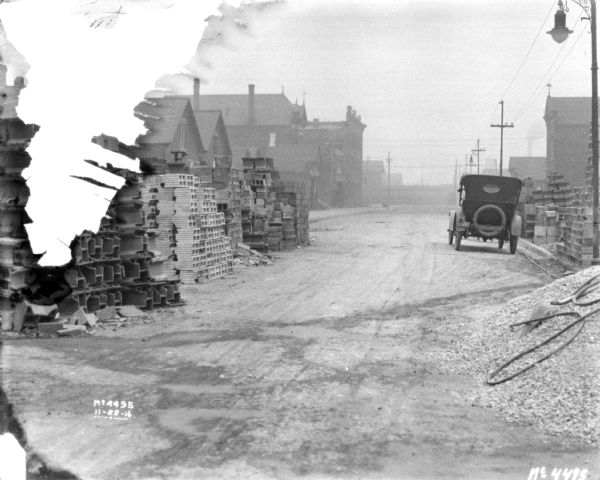 Bricks are stacked along the left side of a road. On the right is a pile of gravel or other material near an automobile. Buildings are further down the road.