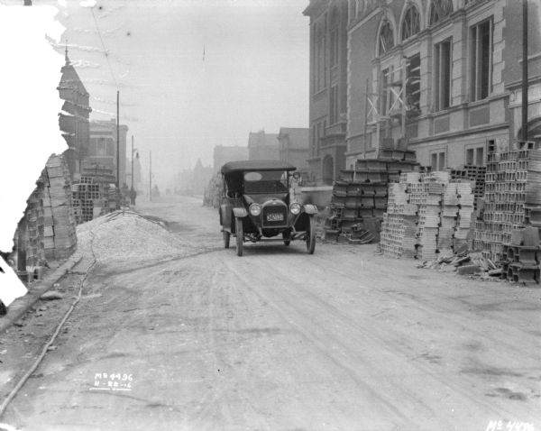 Bricks are stacked along both sides of a road. An automobile is in the center of the road, and a large building is on the right.