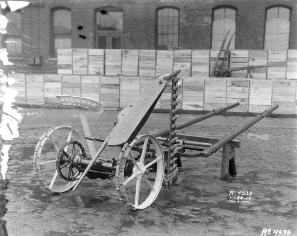 Three-quarter view from right rear of a Milwaukee Mower outdoors in the factory yard. A sign on the Mower blade reads "Milwaukee" with a sub-caption in French below. Crates, probably for shipping, are stacked near a brick building in the background.