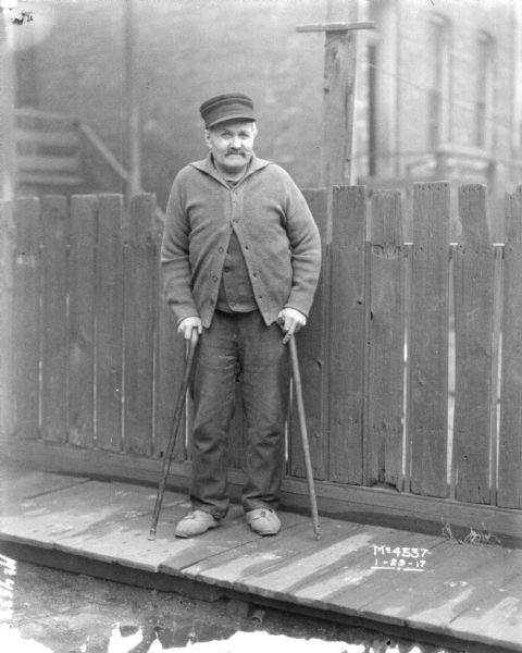 Elderly man with two crutches posing standing on a wood sidewalk in front of a fence.