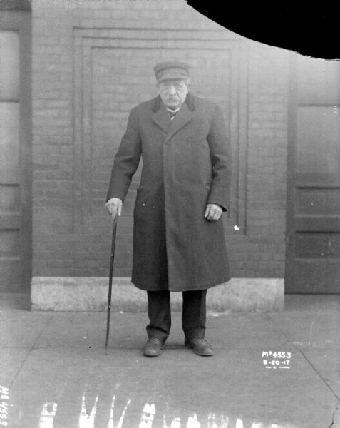 Full-length portrait of an elderly man, in warm dress coat, standing with cane on stairs in front of a brick building.