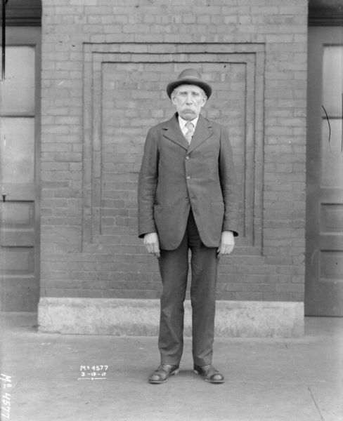 Full-length portrait of a man wearing a suit and hat standing in front of a brick building.