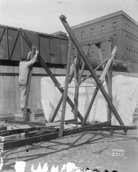 Man standing on a box and reaching up while attaching parts to a Hay Stacker outdoors in the factory yard. Railroad cars are on an elevated platform in the background.