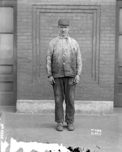 Full-length portrait of a man standing in front of a brick factory wall outdoors.