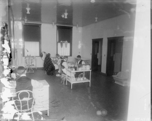Interior view of doctors treatment room. A man is sitting at a desk with his back to the camera on the left man. In the middle of the room one man is treating a man, and another man is treating a woman.