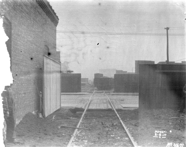 View down set of railroad tracks that cross a street through an open gate and head into a railroad yard. There is a man standing near a brick building on the left near a pile of dirt, and another man is standing near a fence on the right.  