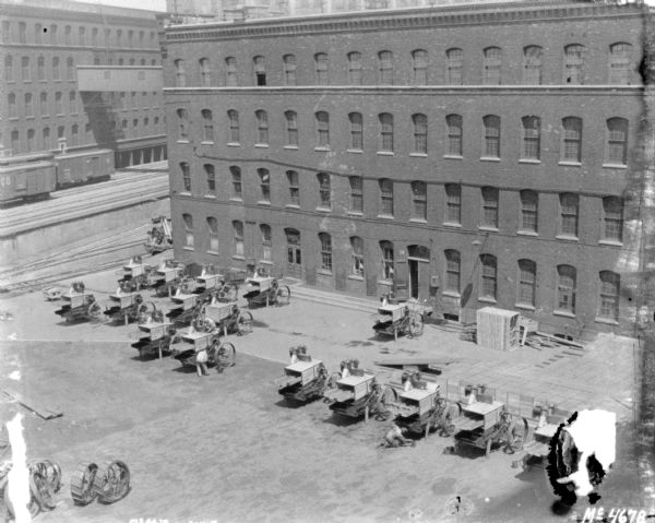 Elevated view of a large group of products, Harvester Threshers, being assembled in the factory yard by men. Other machines are crated nearby. Railroad cars are on an elevated platform between two factory buildings. A skybridge connects the two buildings above the railroad tracks.