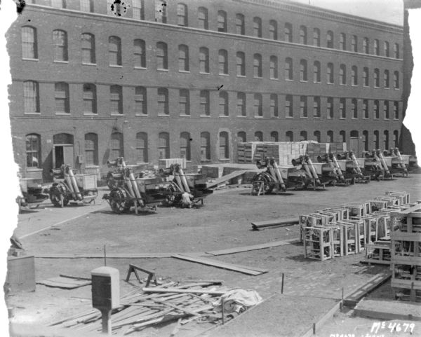 Slightly elevated view of a large group of products, Harvester Threshers, being assembled in the factory yard by men. Other machines are crated nearby, and a brick factory building is in the background.