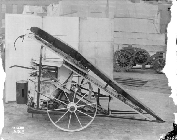Corn Binder in factory yard. In the background are backdrops, a man standing on the left, and brick factory buildings. A tractor is in the background on the right.