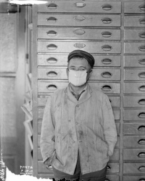 Young man posing standing in front of a chest of drawers. He has a cloth tied over his mouth and nose.