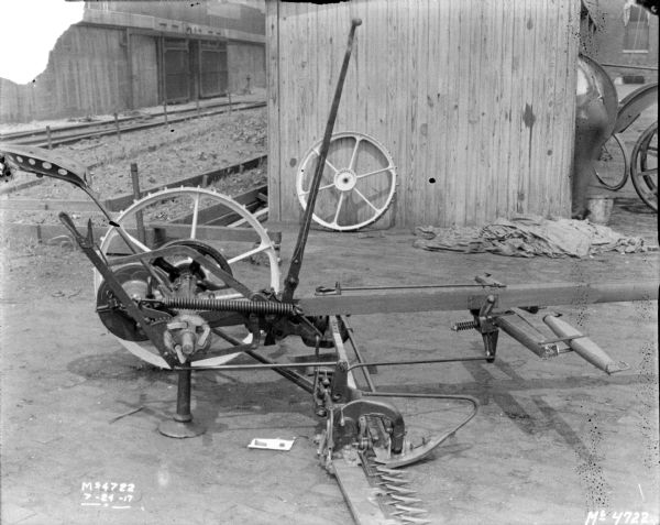 Rear view of a mower parked near a shed in factory yard. The front of a tractor is protruding out from the shed, and a handcart is nearby. Brick buildings are in the background.