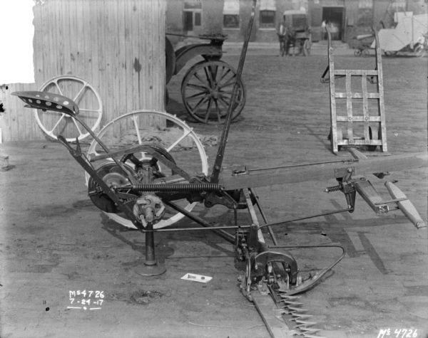 Right side view of a mower parked near a shed in factory yard. The front of a tractor is protruding out from the shed, and a handcart is nearby. Brick buildings and a horse-drawn wagon are in the background.