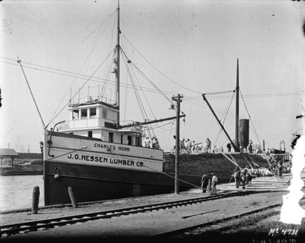 View across railroad tracks towards a ship, with the name: "Charles Horn, J.O. Nessen Lumber Co." on the side. There are buildings and railroad cars on the opposite shoreline. A large group of men are unloading lumber from the heavily loaded ship.
