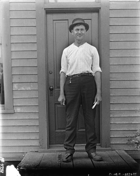 Full-length portrait of a man standing on the stoop in front of a wood sided building. He is holding a rolled up paper in his left hand.