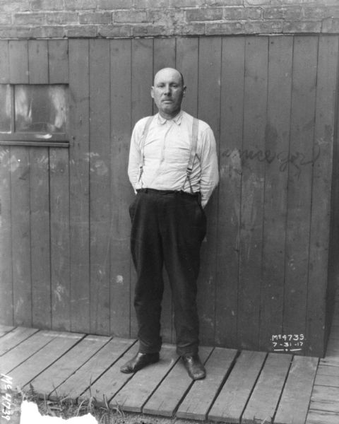 Full-length portrait of a man wearing work clothes standing on a wooden sidewalk in front of a wood wall, with a brick wall above.