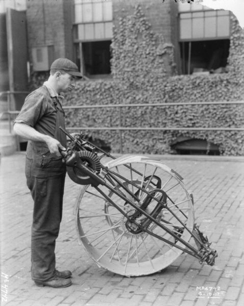 Employee standing and holding an agricultural machinery part, including a large wheel, in factory yard. A brick building is in the background.