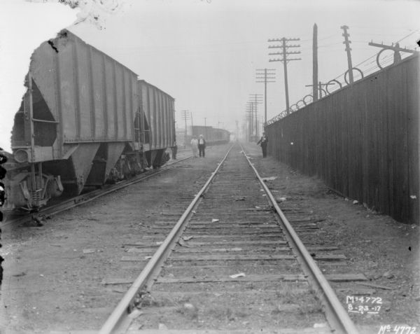 View down railroad tracks. There are railroad cars on the left on another set of tracks. Men are standing further down the tracks, and a man standing near a tall fence on the right is pointing down at the tracks. There is barbed wire on top of the fence. Power lines are in the background.