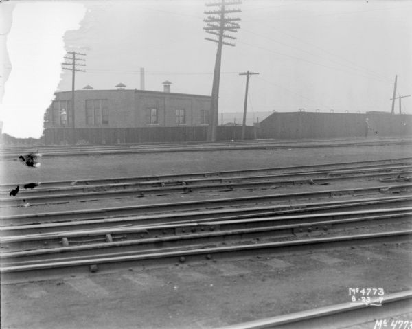 View across multiple sets of railroad tracks towards a factory building. A man is standing on the railroad tracks in the background on the right in front of a tall fence with barbed wire on top.