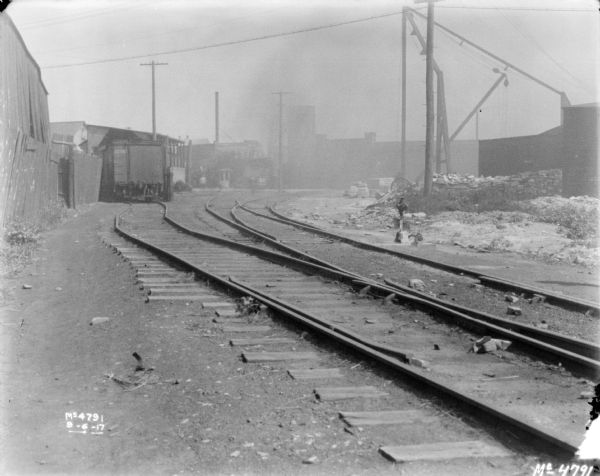 View down side of railroad tracks, wit a fence along the left. A railroad car is further down the tracks near buildings.